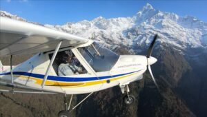 Ultralight Flight in Pokhara-An orange and white ultralight aircraft with registration 9N-AKJ flying over green mountain valleys and snow-capped mountains.