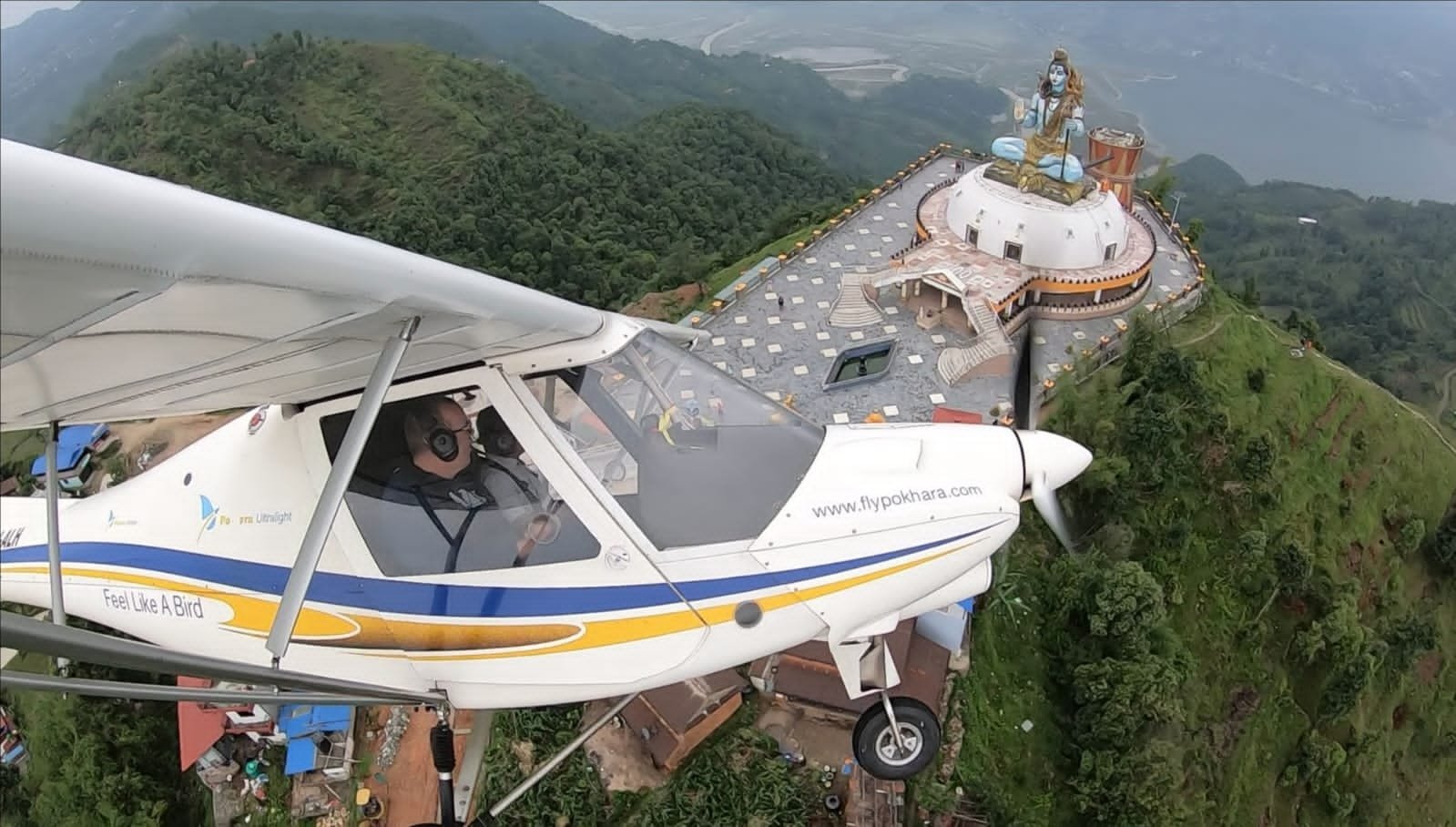 Ultralight Flight in Pokhara-A white light aircraft flying close to a large white platform featuring a giant statue of Lord Shiva overlooking green hills and a lake.