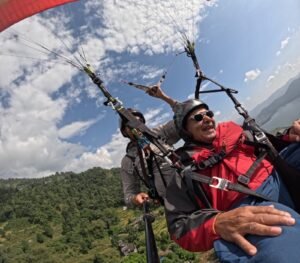 Paragliding in Pokhara-Two men tandem paragliding high above green hills, a large lake, and scattered houses under a partly cloudy sky, holding a selfie stick.