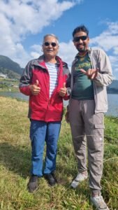 Paragliding in Pokhara-Two men standing side by side outdoors on grassy ground near a lake, wearing sunglasses and jackets, smiling and gesturing with their hands.
