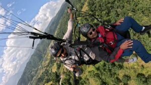 Paragliding in Pokhara-Two men standing side by side on grassy terrain near a lake, wearing jackets and sunglasses, giving thumbs-up and hand gestures.
